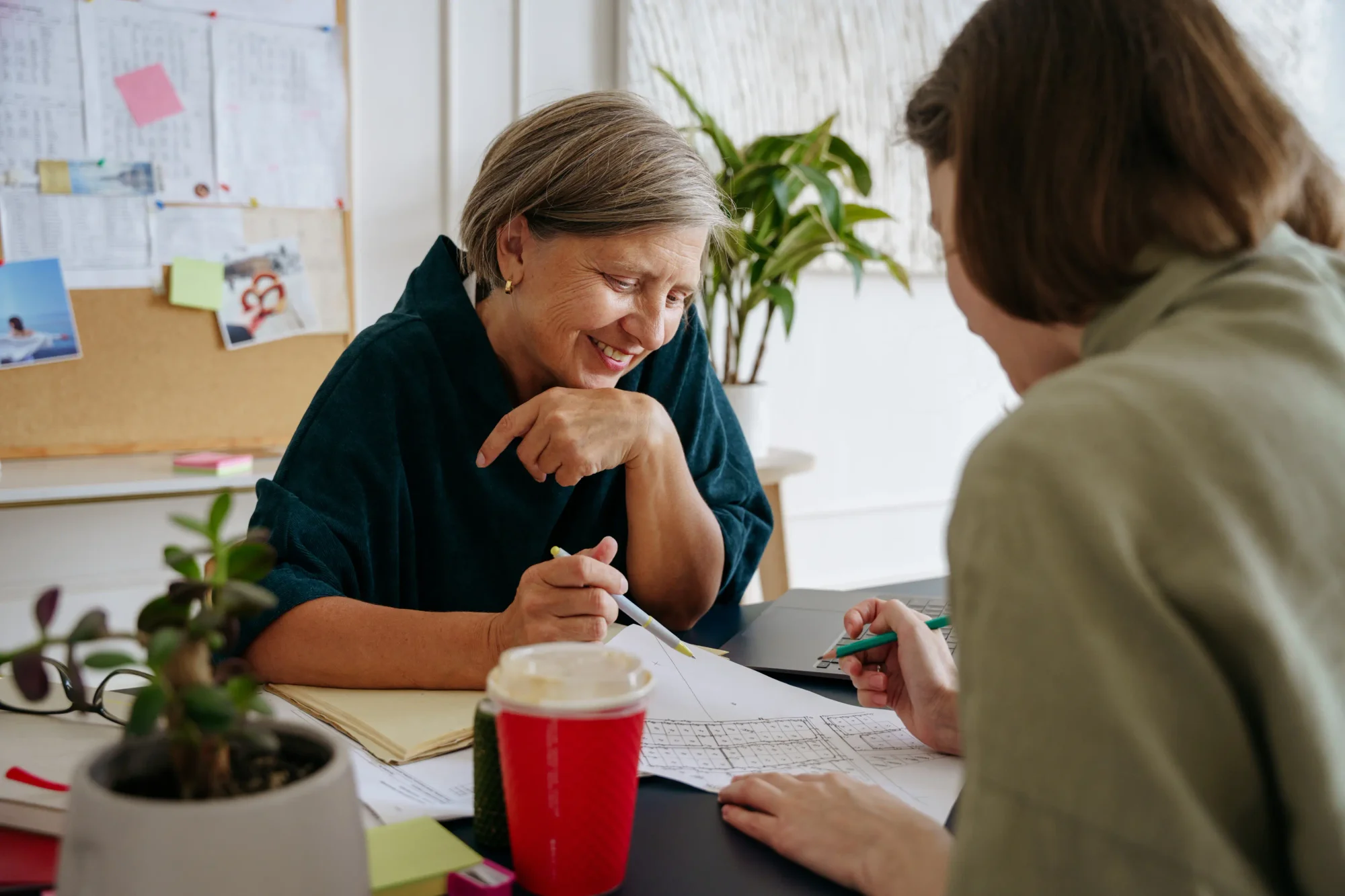 Two women are engaged in a discussion at a table, with one smiling and pointing at a document while the other writes, surrounded by plants and notes in a bright workspace