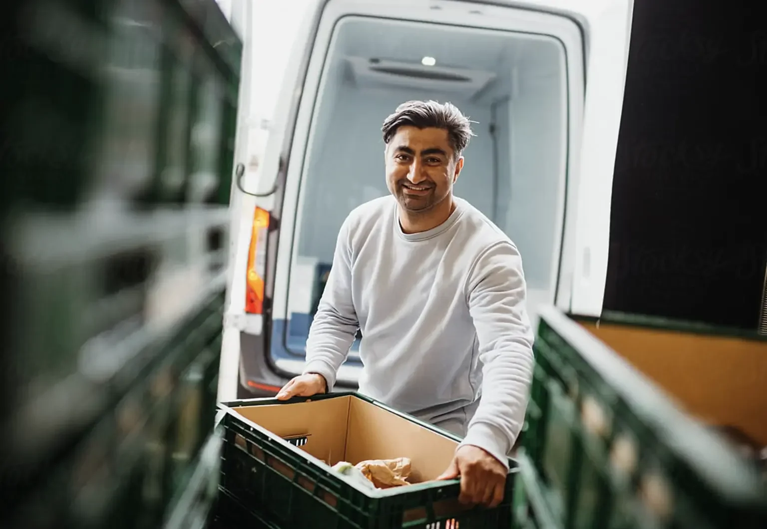 A smiling man in a light gray sweatshirt carries a box of goods while standing next to a delivery van filled with green crates