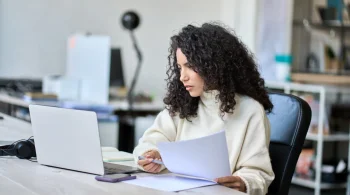 A woman sits at her desk and reviews documents on her laptop.